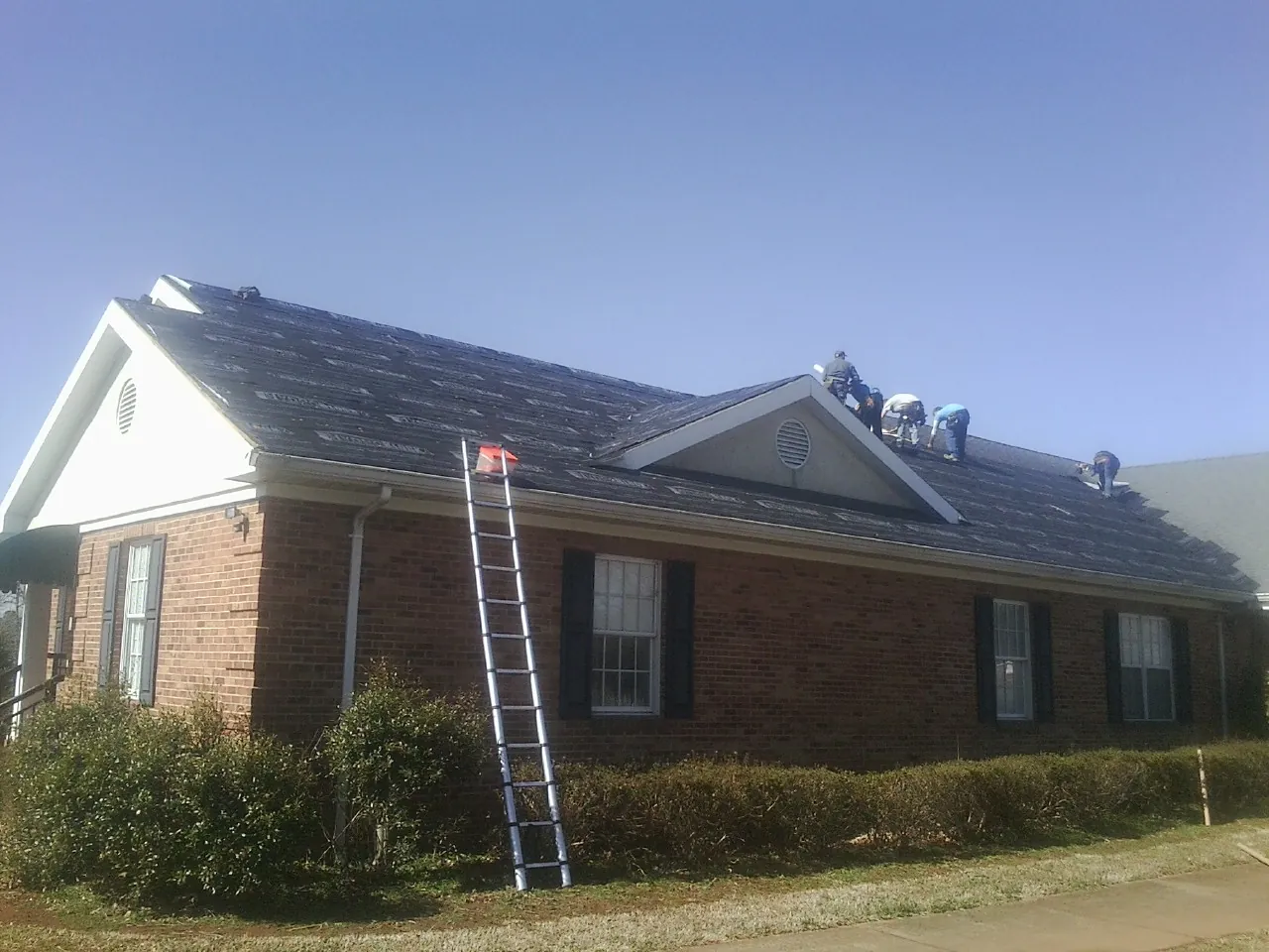 Crew tearing shingles off a brick ranch roof with a ladder set — Emergency Roof Repair work by our crew in Altona